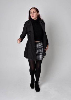 Siyah ceket, tartan etek ve çizme giyen kızıl saçlı bir kızın tam boy portresi. Stüdyo geçmişine karşı poz vermek.