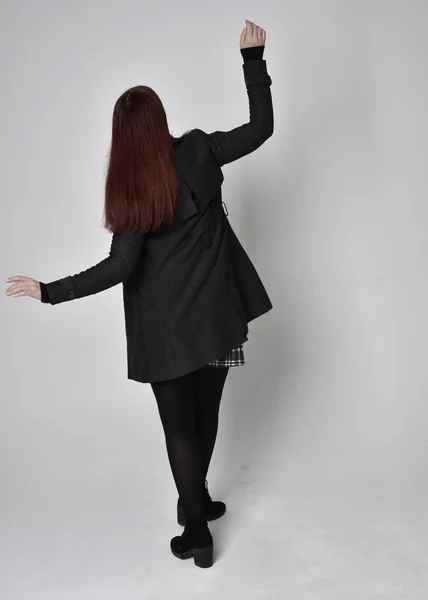 黒いコート タータンスカートとブーツを身に着けている赤い髪の少女の完全な長さの肖像画 撮影所を背景にカメラに背を向けて立ちポーズ — ストック写真