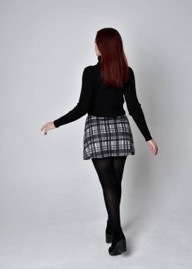 Siyah balıkçı yaka kazak, ekose etek ve çizme giyen kızıl saçlı bir kızın tam boy portresi. Stüdyonun arka planında kameraya dönük poz vermek.