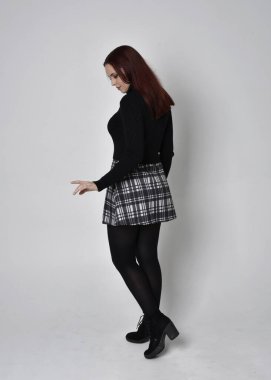 Siyah balıkçı yaka kazak, ekose etek ve çizme giyen kızıl saçlı bir kızın tam boy portresi. Stüdyonun arka planında kameraya dönük poz vermek.