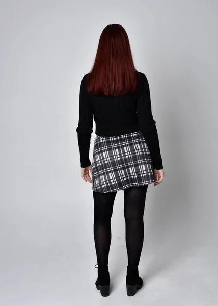 黒のタートルネックセーターを着て 赤い髪の少女の完全な長さの肖像画は スカートとブーツを果たした 撮影所を背景にカメラに背を向けて立ちポーズ — ストック写真