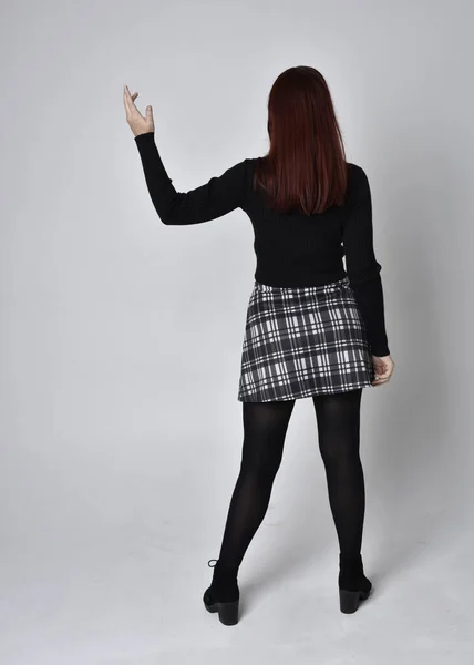 全长肖像一个红头发的女孩穿着黑色高领毛衣 格子裙和靴子 在摄影棚的背景下 背对着相机站姿 — 图库照片