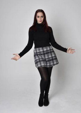 Full length portrait of a red haired  girl wearing black turtleneck sweater,  plaid skirt and boots.  Standing pose against a studio background. clipart