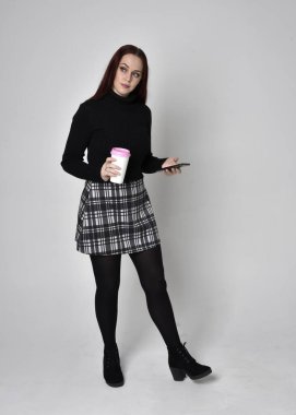 Siyah balıkçı yaka kazak, ekose etek ve çizme giyen kızıl saçlı bir kızın tam boy portresi. Stüdyo geçmişine karşı poz vermek.