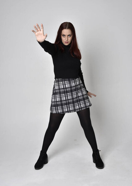 Портрет рыжеволосой девушки в черном свитере в водолазке, клетчатой юбке и сапогах. Стоя позируют на студийном фоне.