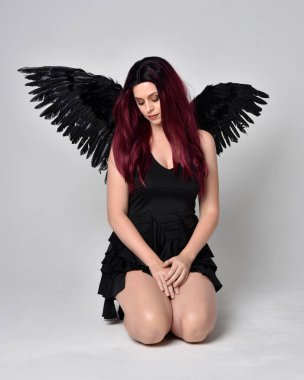 Siyah elbise ve tüylü melek kanatları takan kızıl saçlı bir kızın tam boy portresi. Stüdyonun arka planında poz vermek.