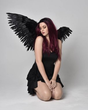 Siyah elbise ve tüylü melek kanatları takan kızıl saçlı bir kızın tam boy portresi. Stüdyonun arka planında poz vermek.