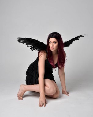 Siyah elbise ve tüylü melek kanatları takan kızıl saçlı bir kızın tam boy portresi. Stüdyonun arka planında poz vermek.