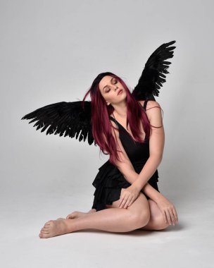 Siyah elbise ve tüylü melek kanatları takan kızıl saçlı bir kızın tam boy portresi. Stüdyonun arka planında poz vermek.