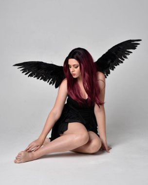Siyah elbise ve tüylü melek kanatları takan kızıl saçlı bir kızın tam boy portresi. Stüdyonun arka planında poz vermek.