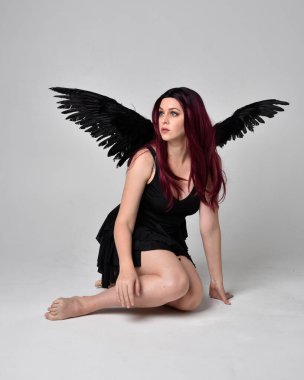 Siyah elbise ve tüylü melek kanatları takan kızıl saçlı bir kızın tam boy portresi. Stüdyonun arka planında poz vermek.