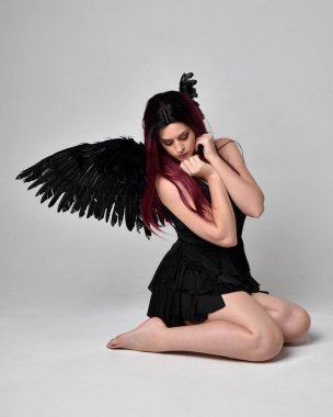 Siyah elbise ve tüylü melek kanatları takan kızıl saçlı bir kızın tam boy portresi. Stüdyonun arka planında poz vermek.