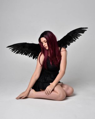 Siyah elbise ve tüylü melek kanatları takan kızıl saçlı bir kızın tam boy portresi. Stüdyonun arka planında poz vermek.