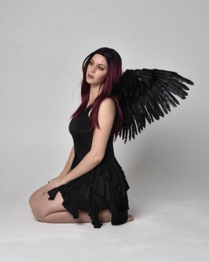 Siyah elbise ve tüylü melek kanatları takan kızıl saçlı bir kızın tam boy portresi. Stüdyonun arka planında poz vermek.