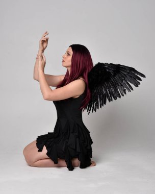 Siyah elbise ve tüylü melek kanatları takan kızıl saçlı bir kızın tam boy portresi. Stüdyonun arka planında poz vermek.