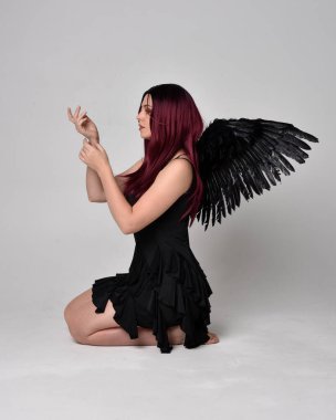 Siyah elbise ve tüylü melek kanatları takan kızıl saçlı bir kızın tam boy portresi. Stüdyonun arka planında poz vermek.