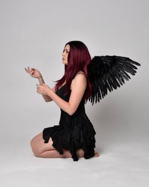Siyah elbise ve tüylü melek kanatları takan kızıl saçlı bir kızın tam boy portresi. Stüdyonun arka planında poz vermek.