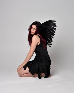Siyah elbise ve tüylü melek kanatları takan kızıl saçlı bir kızın tam boy portresi. Stüdyonun arka planında poz vermek.