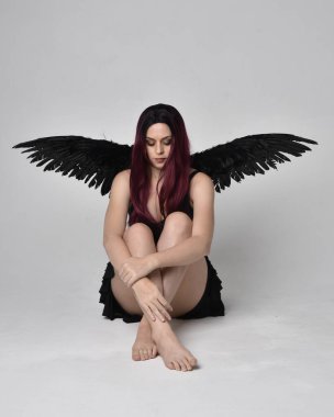 Siyah elbise ve tüylü melek kanatları takan kızıl saçlı bir kızın tam boy portresi. Stüdyonun arka planında poz vermek.