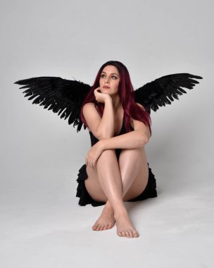 Siyah elbise ve tüylü melek kanatları takan kızıl saçlı bir kızın tam boy portresi. Stüdyonun arka planında poz vermek.