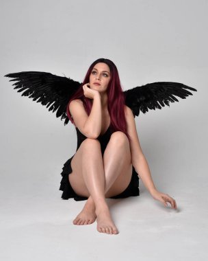 Siyah elbise ve tüylü melek kanatları takan kızıl saçlı bir kızın tam boy portresi. Stüdyonun arka planında poz vermek.