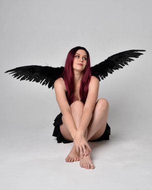 Siyah elbise ve tüylü melek kanatları takan kızıl saçlı bir kızın tam boy portresi. Stüdyonun arka planında poz vermek.