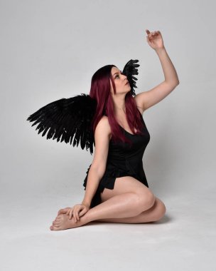 Siyah elbise ve tüylü melek kanatları takan kızıl saçlı bir kızın tam boy portresi. Stüdyonun arka planında poz vermek.