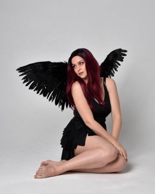 Siyah elbise ve tüylü melek kanatları takan kızıl saçlı bir kızın tam boy portresi. Stüdyonun arka planında poz vermek.