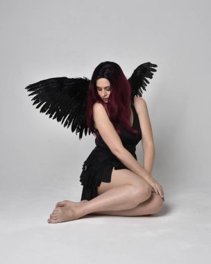 Siyah elbise ve tüylü melek kanatları takan kızıl saçlı bir kızın tam boy portresi. Stüdyonun arka planında poz vermek.