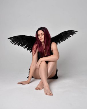Siyah elbise ve tüylü melek kanatları takan kızıl saçlı bir kızın tam boy portresi. Stüdyonun arka planında poz vermek.