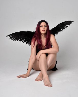 Siyah elbise ve tüylü melek kanatları takan kızıl saçlı bir kızın tam boy portresi. Stüdyonun arka planında poz vermek.