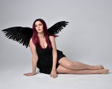 Siyah elbise ve tüylü melek kanatları takan kızıl saçlı bir kızın tam boy portresi. Stüdyonun arka planında poz vermek.