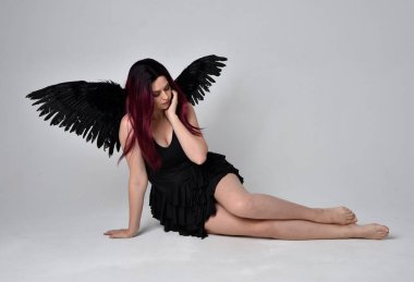 Siyah elbise ve tüylü melek kanatları takan kızıl saçlı bir kızın tam boy portresi. Stüdyonun arka planında poz vermek.