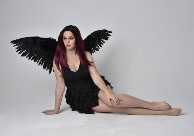 Siyah elbise ve tüylü melek kanatları takan kızıl saçlı bir kızın tam boy portresi. Stüdyonun arka planında poz vermek.