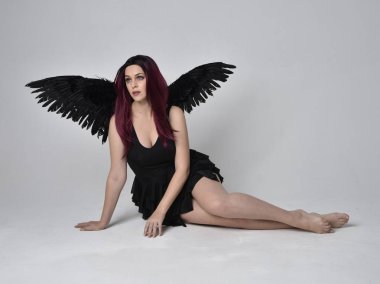 Siyah elbise ve tüylü melek kanatları takan kızıl saçlı bir kızın tam boy portresi. Stüdyonun arka planında poz vermek.