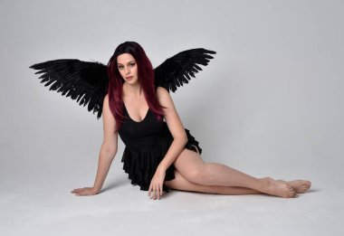 Siyah elbise ve tüylü melek kanatları takan kızıl saçlı bir kızın tam boy portresi. Stüdyonun arka planında poz vermek.