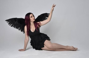 Siyah elbise ve tüylü melek kanatları takan kızıl saçlı bir kızın tam boy portresi. Stüdyonun arka planında poz vermek.