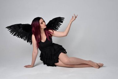 Siyah elbise ve tüylü melek kanatları takan kızıl saçlı bir kızın tam boy portresi. Stüdyonun arka planında poz vermek.