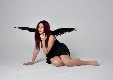 Siyah elbise ve tüylü melek kanatları takan kızıl saçlı bir kızın tam boy portresi. Stüdyonun arka planında poz vermek.