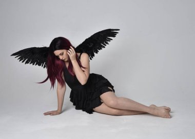 Siyah elbise ve tüylü melek kanatları takan kızıl saçlı bir kızın tam boy portresi. Stüdyonun arka planında poz vermek.
