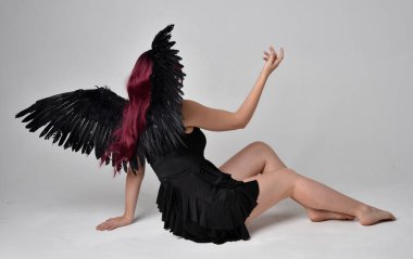 Siyah elbise ve tüylü melek kanatları takan kızıl saçlı bir kızın tam boy portresi. Stüdyonun arka planında poz vermek.