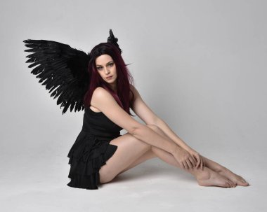Siyah elbise ve tüylü melek kanatları takan kızıl saçlı bir kızın tam boy portresi. Stüdyonun arka planında poz vermek.