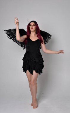 Siyah elbise ve tüylü melek kanatları takan kızıl saçlı bir kızın tam boy portresi. Stüdyo geçmişine karşı poz vermek.