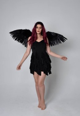 Siyah elbise ve tüylü melek kanatları takan kızıl saçlı bir kızın tam boy portresi. Stüdyo geçmişine karşı poz vermek.