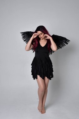 Siyah elbise ve tüylü melek kanatları takan kızıl saçlı bir kızın tam boy portresi. Stüdyo geçmişine karşı poz vermek.