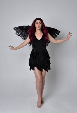 Siyah elbise ve tüylü melek kanatları takan kızıl saçlı bir kızın tam boy portresi. Stüdyo geçmişine karşı poz vermek.