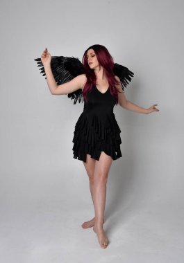 Siyah elbise ve tüylü melek kanatları takan kızıl saçlı bir kızın tam boy portresi. Stüdyo geçmişine karşı poz vermek.