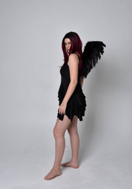 Siyah elbise ve tüylü melek kanatları takan kızıl saçlı bir kızın tam boy portresi. Stüdyo geçmişine karşı poz vermek.