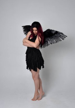 Siyah elbise ve tüylü melek kanatları takan kızıl saçlı bir kızın tam boy portresi. Stüdyo geçmişine karşı poz vermek.