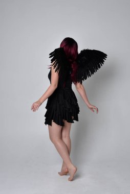 Siyah elbise ve tüylü melek kanatları takan kızıl saçlı bir kızın tam boy portresi. Stüdyo geçmişine karşı poz vermek.